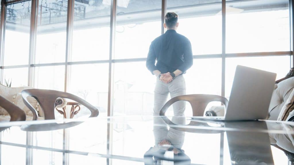 A man stands facing large bright windows in a modern room, his hands behind his back.
