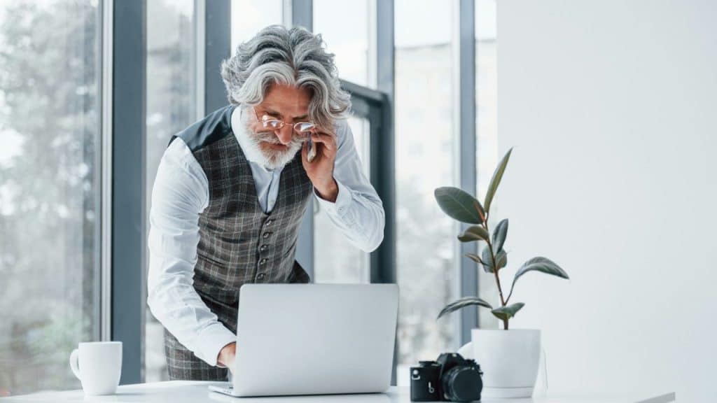 An older man in a vest and glasses works on a laptop at a desk near a window.