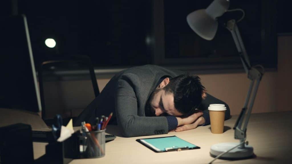 A man in a suit is asleep face down on an office desk late at night.