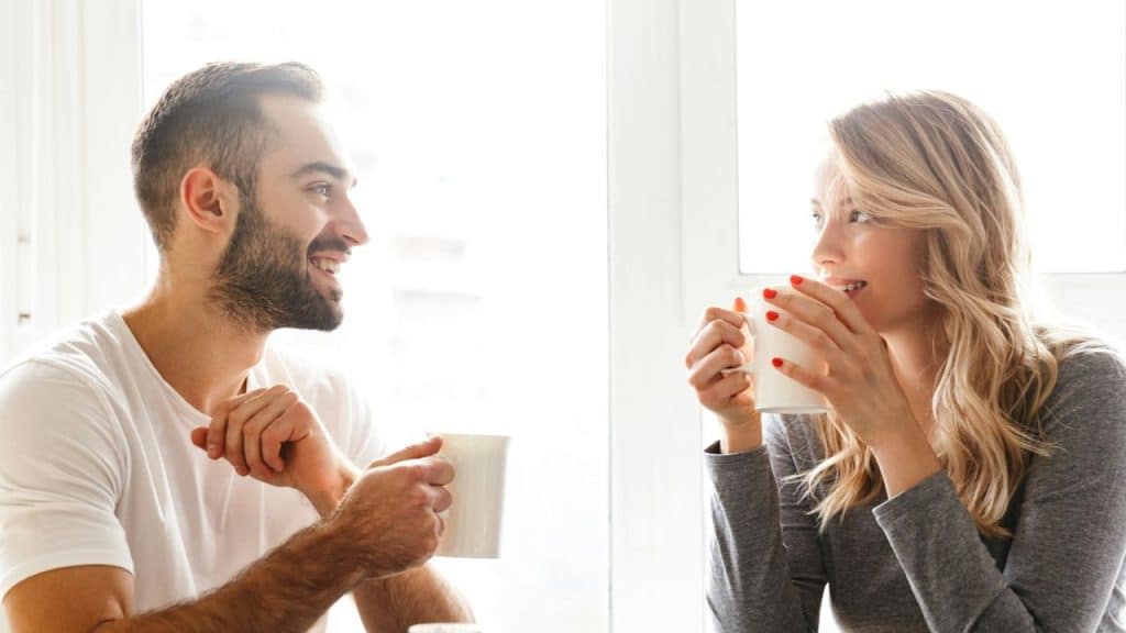 Smiling couple sitting across from each other, each holding a white mug near a bright window.