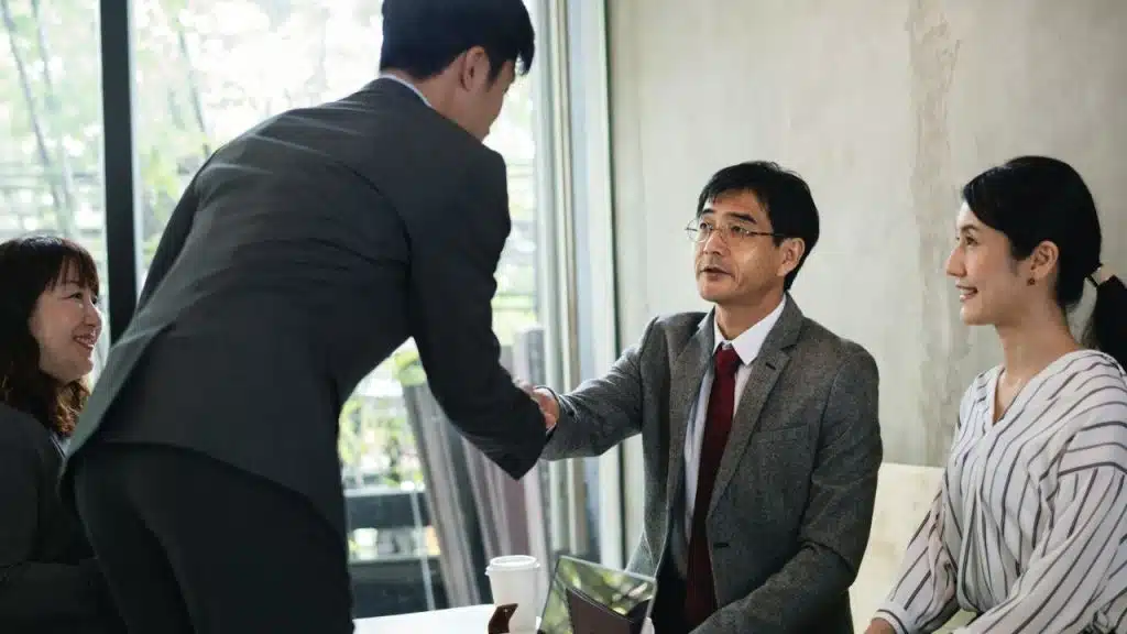 An Asian businessman in a suit shakes hands with another man during an office meeting or interview.