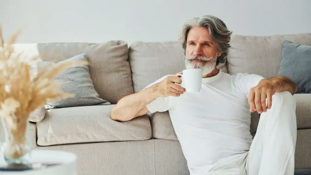 Older man with white hair and beard, holding a mug, leans against a light gray couch.