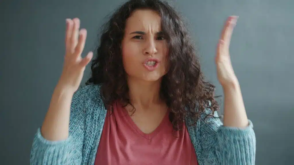 Woman with curly hair scowling and gesturing emphatically against a plain gray background.