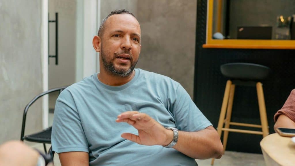 Bearded man in a light blue T-shirt talking and gesturing with his hand in an indoor cafe.