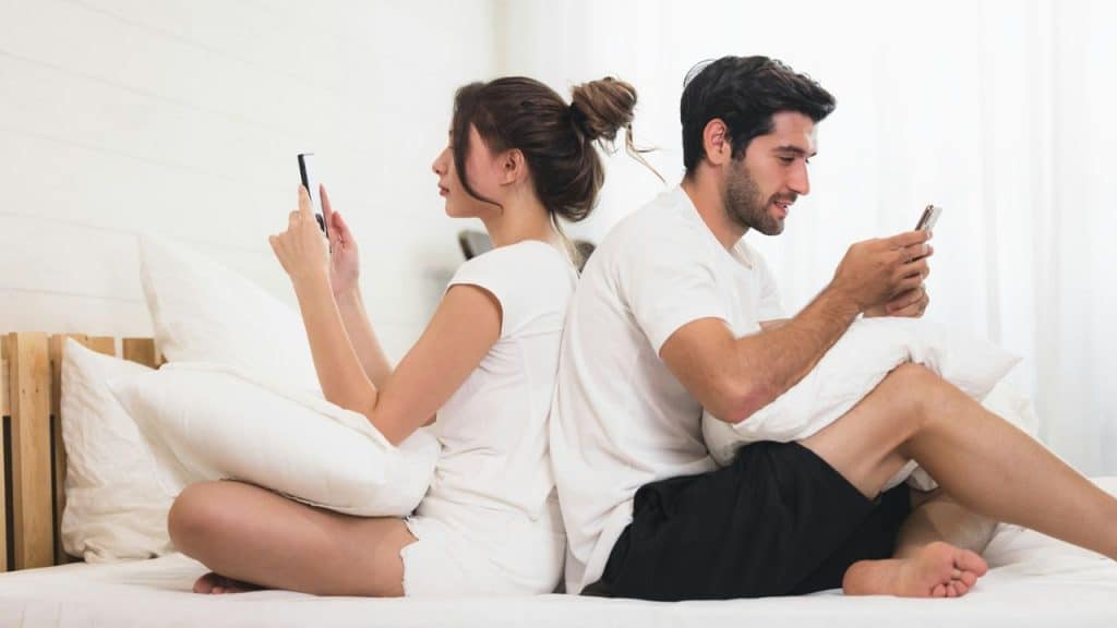 A man and woman sit back-to-back in bed, each intently looking at their phone.