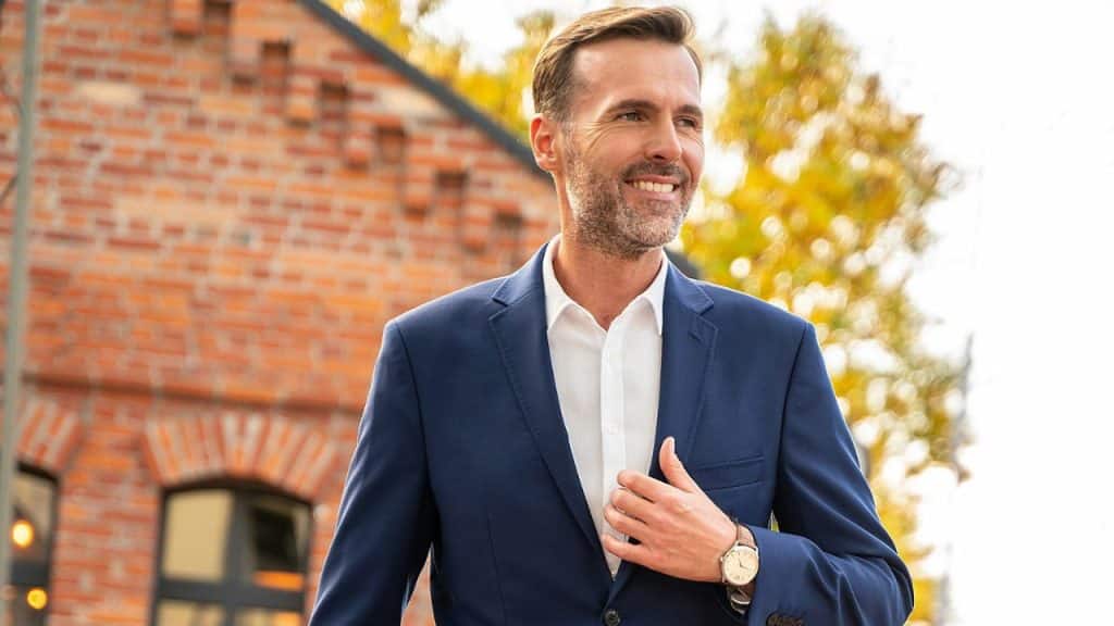 A man in a navy suit jacket and white shirt smiling outdoors next to a brick building.