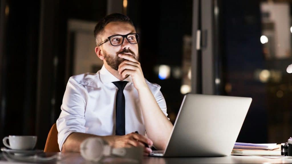 Man in a white shirt and tie sitting at a desk in the dark, looking thoughtfully away from his laptop.