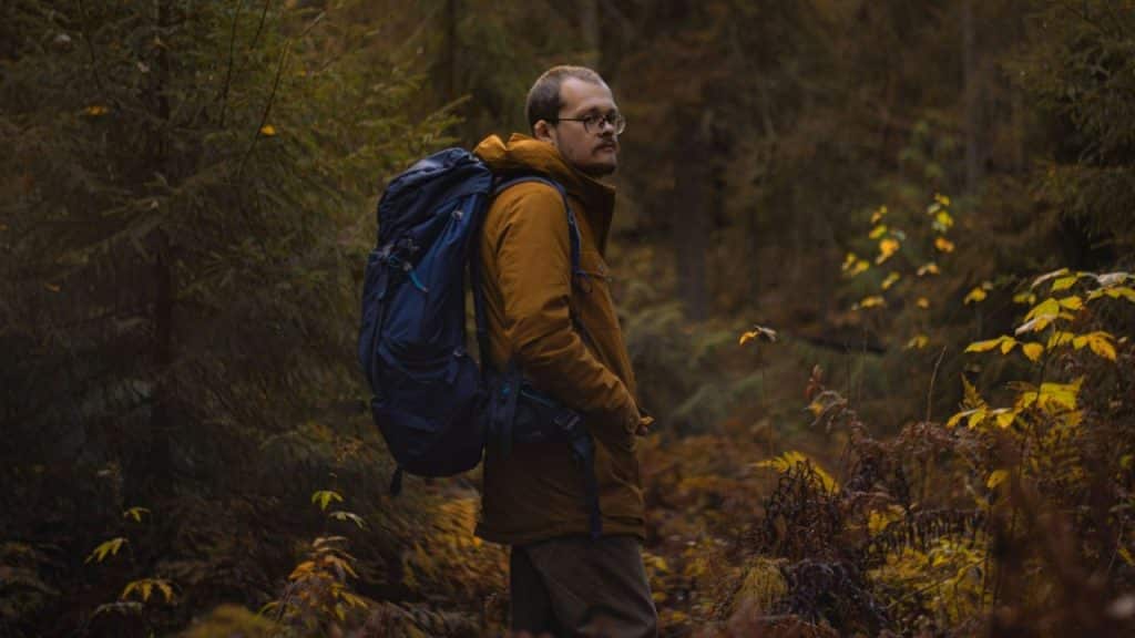 Man wearing a large blue backpack and brown jacket stands in a dense forest.
