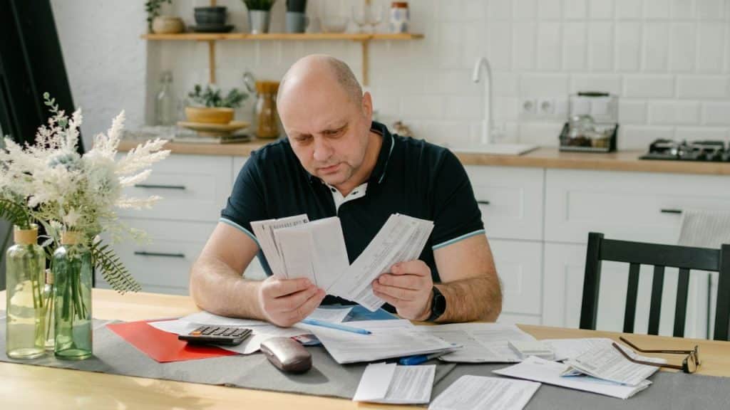 A man in a plaid shirt sits at a table writing with a pencil and calculator nearby.