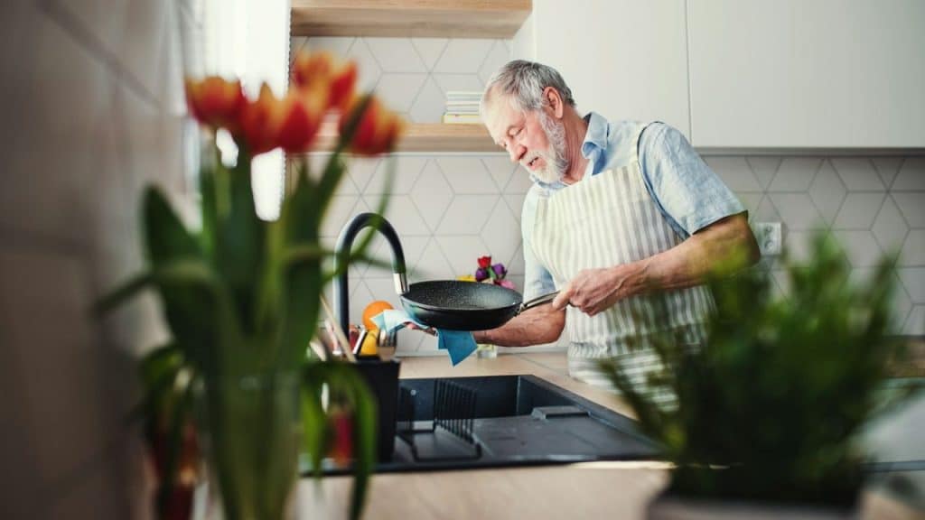 Older man with a beard, wearing a striped apron, washing a frying pan in a kitchen sink.