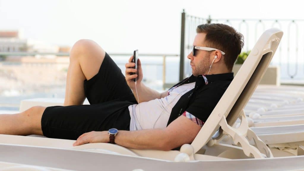 Man in sunglasses and shorts relaxing on a lounge chair, looking at a phone.
