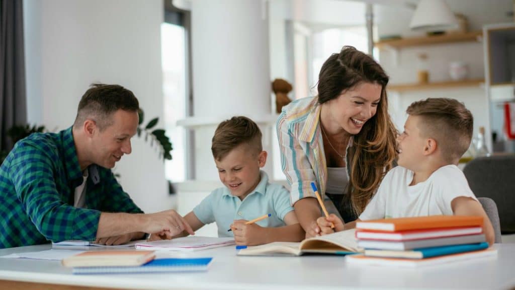 Two smiling adults help two young boys with schoolwork at a brightly lit table.