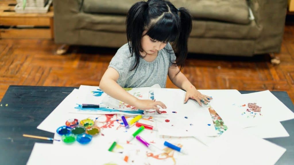 Young girl with pigtails painting with her fingers on paper at a table indoors.