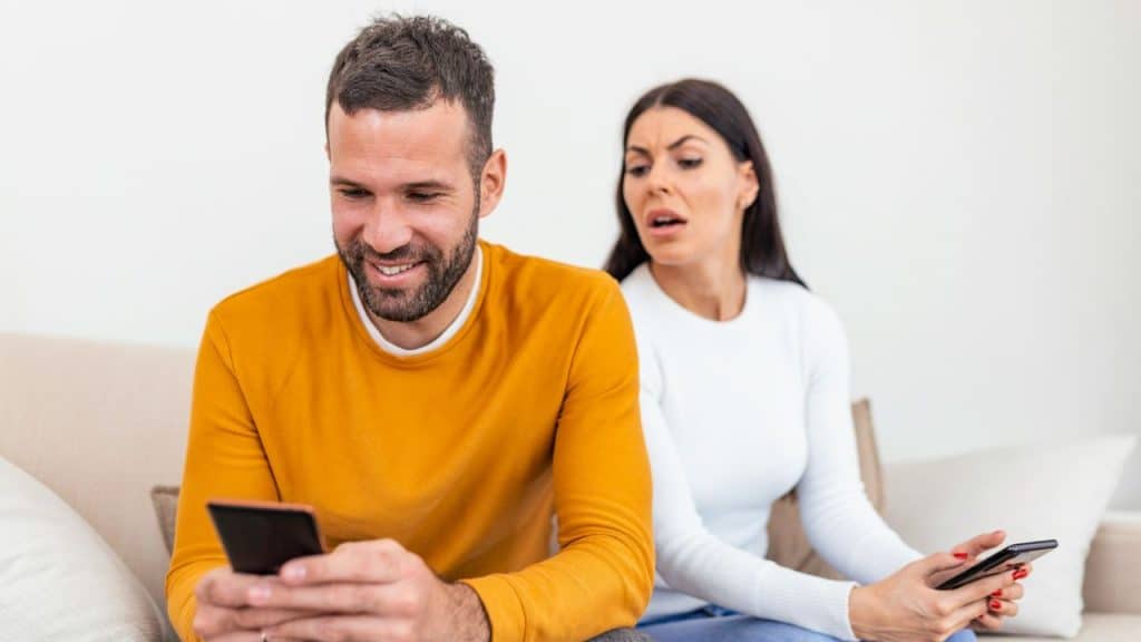 A smiling man is looking at his phone while a suspicious woman peers over his shoulder.