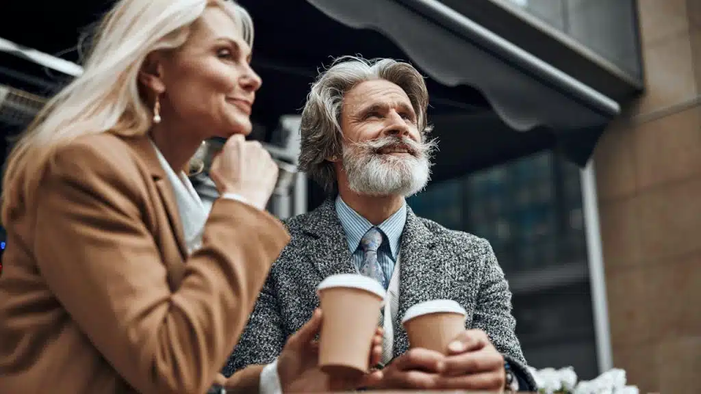 A stylish, mature couple sitting at an outdoor table, smiling and looking up, both holding takeout coffee cups.
