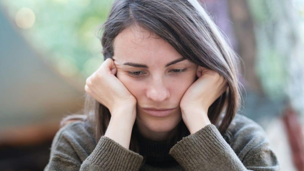Close-up of a young woman with dark hair resting her chin on her hands, looking down with a sad expression.