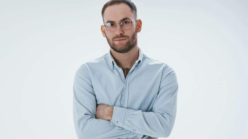 A bearded man wearing a light blue button-down shirt and glasses stands with his arms crossed against a white background.