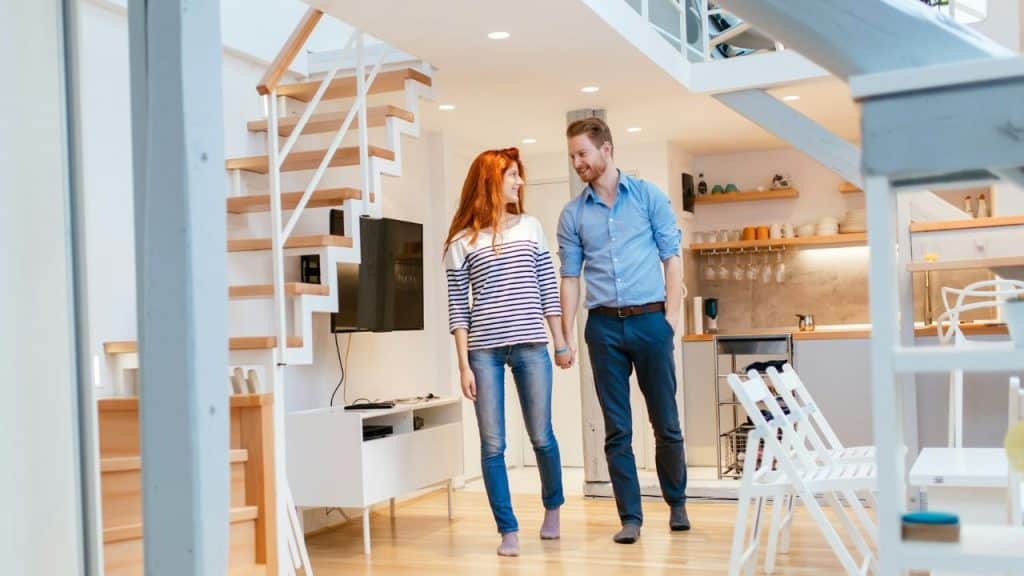 A smiling redheaded woman and a man in a blue shirt hold hands while walking through a bright, modern home.