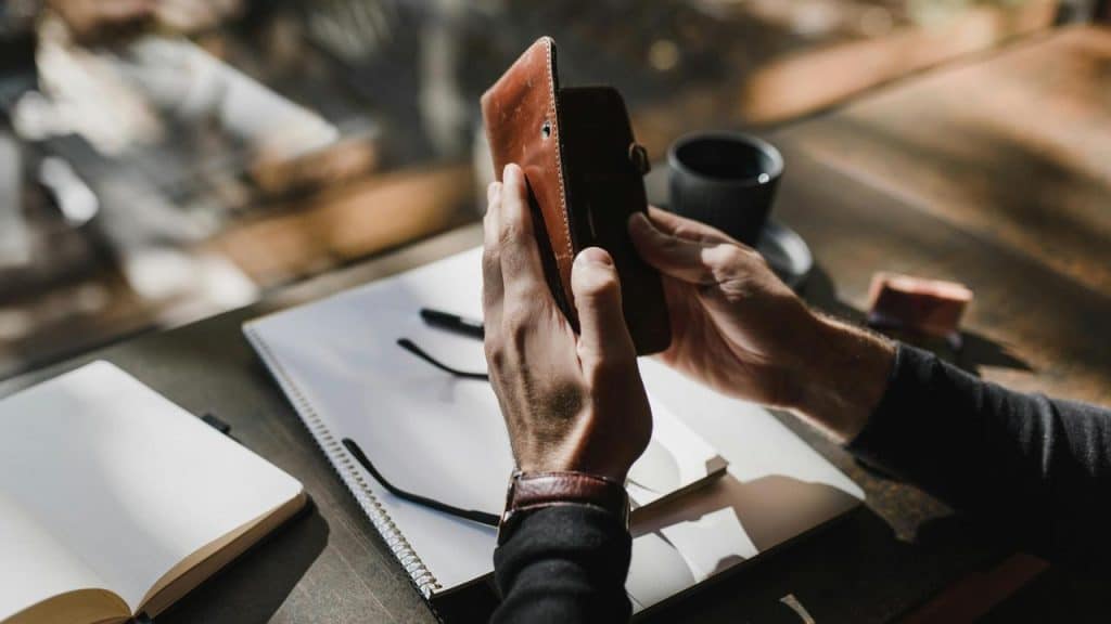 A person holding a brown leather wallet at a desk with notebooks and a coffee cup.
