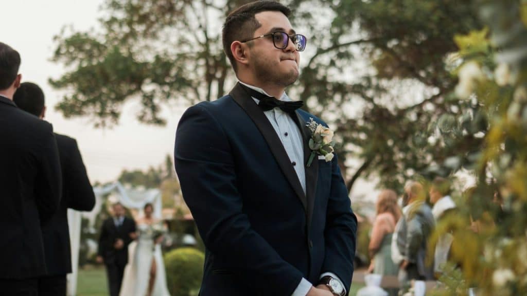 A groom in a dark tuxedo and glasses stands in an outdoor wedding venue with the bride in the blurred background.