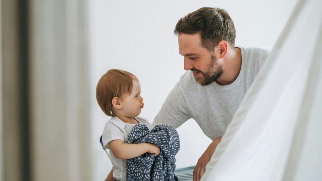 Father in a grey shirt smiling at a young child holding a blanket.
