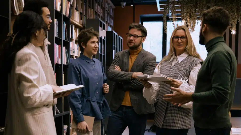 Six adults are standing in a group, talking and holding books, near a bookshelf.