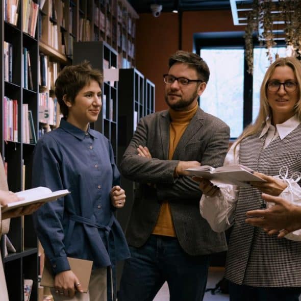 Six adults are standing in a group, talking and holding books, near a bookshelf.