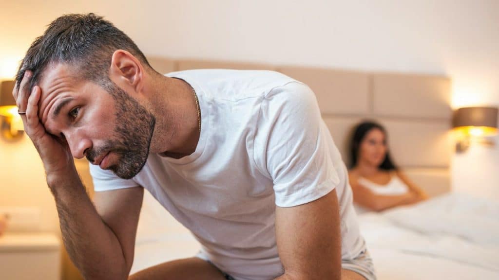 A distraught man in a white t-shirt sits on a bed with his hand on his head, and a woman is blurred in the background.