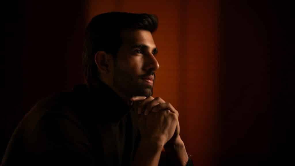A man in a dark turtleneck is sitting in a dimly lit room, with his hands clasped under his chin.