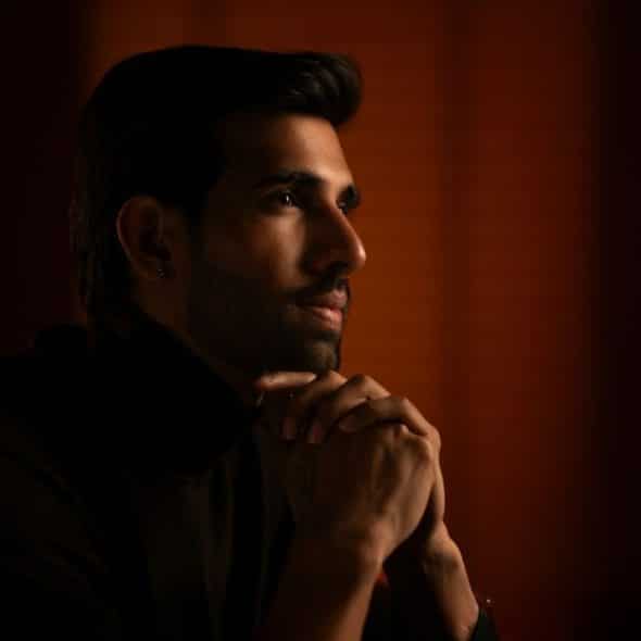 A man in a dark turtleneck is sitting in a dimly lit room, with his hands clasped under his chin.