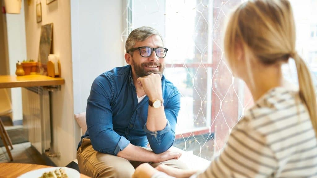 Smiling man with a beard and glasses sits, chin on his hand, facing a woman.