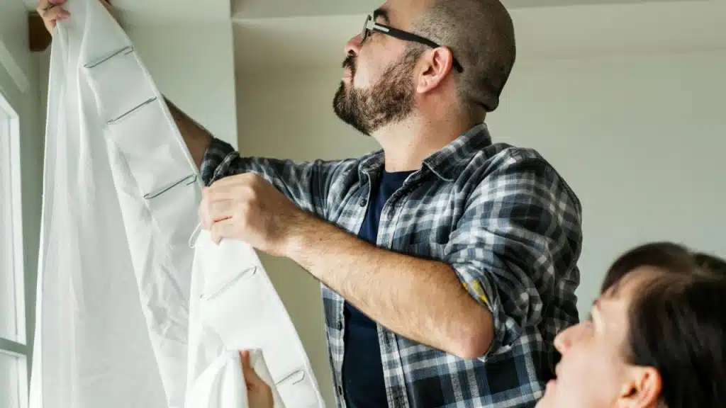 Man in glasses and a plaid shirt hangs a white curtain while a woman looks up at him.