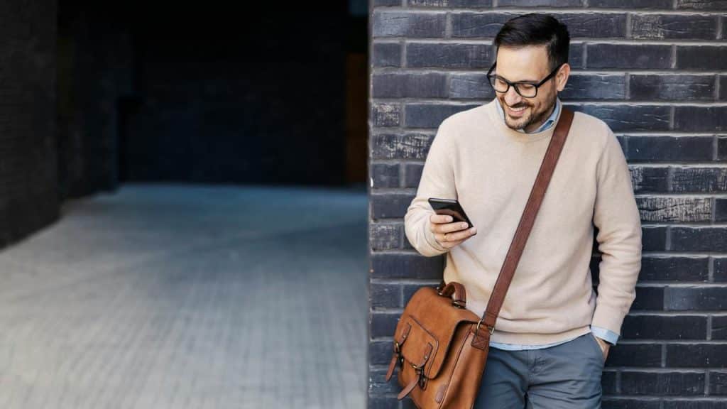 A man wearing glasses and a beige sweater smiling while looking at his phone.