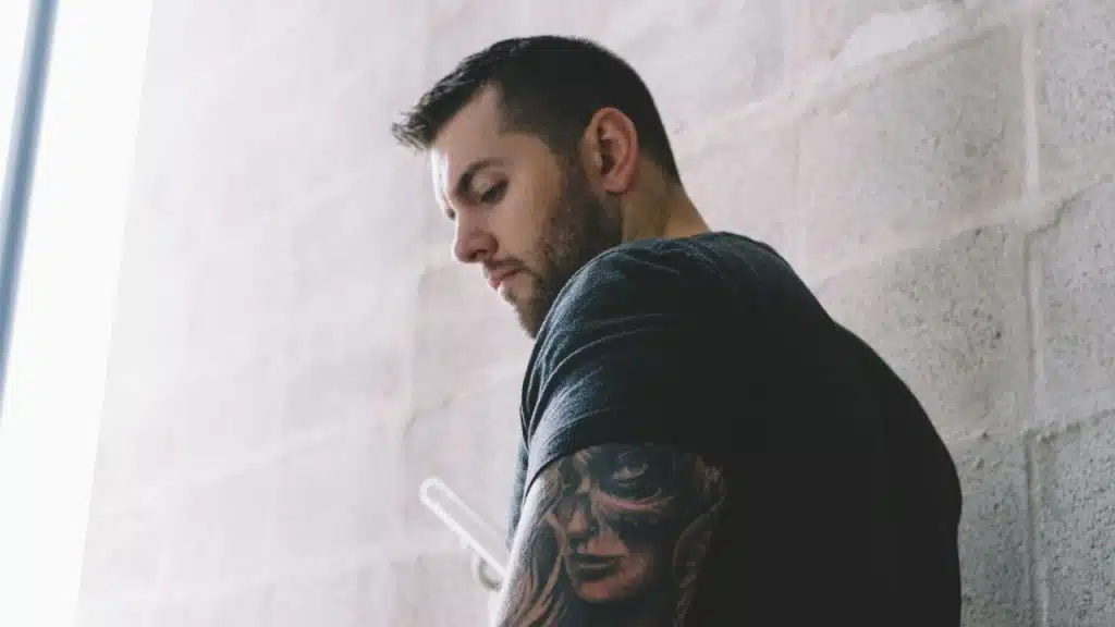 Man with a dark t-shirt and full sleeve tattoo looking down against a brick wall.
