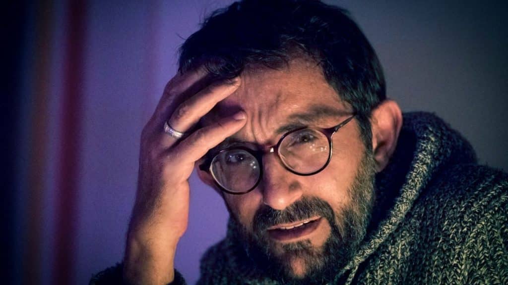 Man in glasses and a sweater with his hand on his forehead, looking confused.