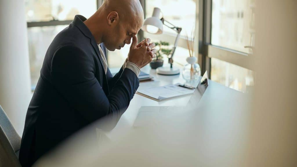 A man in an office suit sitting at a desk with his head bowed and hands together.