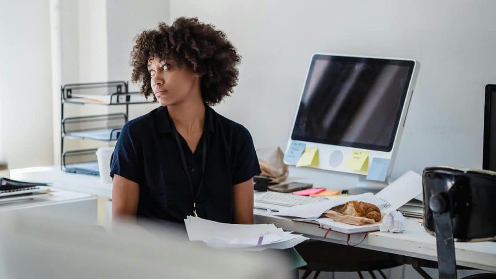 A woman sitting at a cluttered desk in an office, looking away thoughtfully.