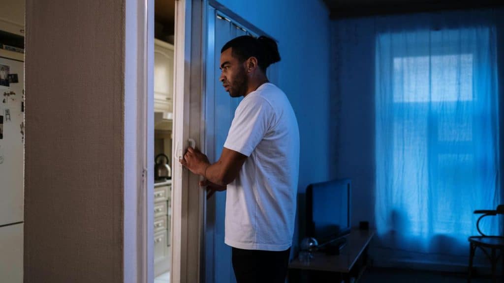 A man standing in a dimly lit room, looking into an open refrigerator at night.
