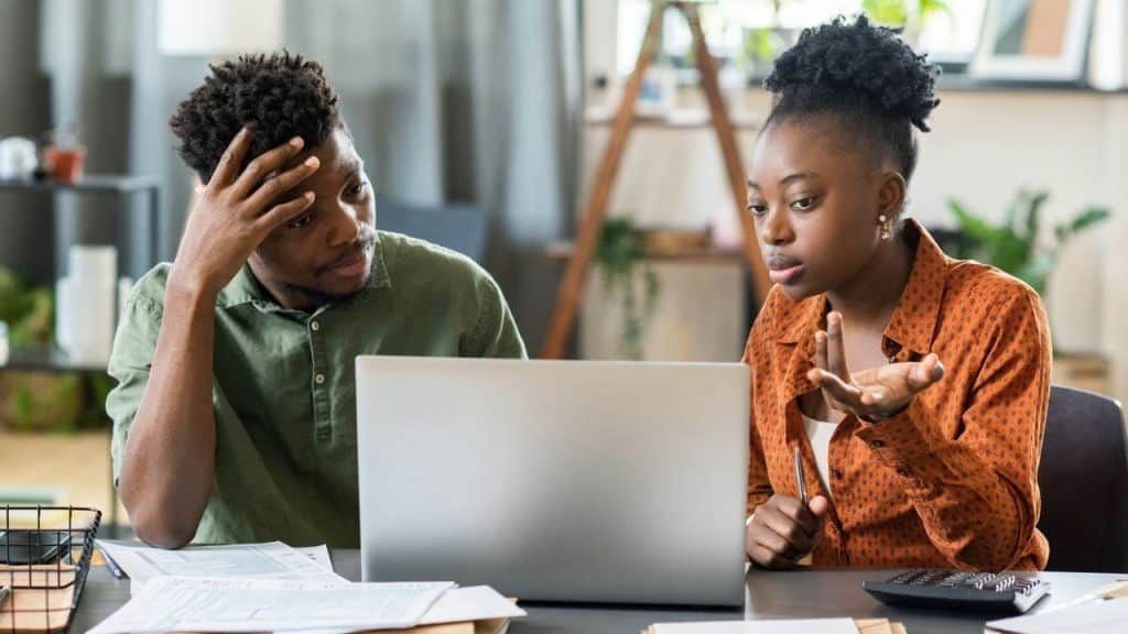 A frustrated man with his hand on his head sits next to a woman who is gesturing while they both look at a laptop.