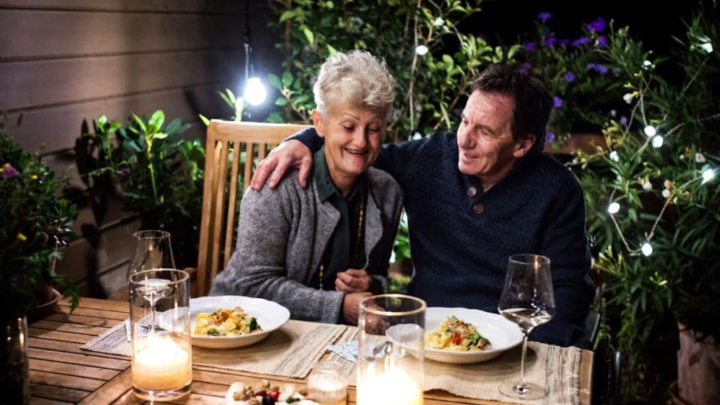 A happy older couple dines outdoors at night, surrounded by string lights and plants.