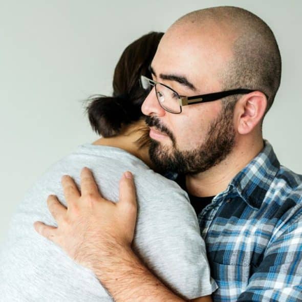 A bald, bearded man in glasses and a plaid shirt hugs a woman.