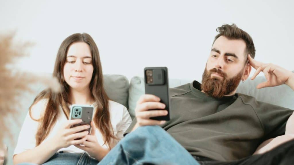 A bearded man and a woman sit on a couch, each looking down at their own smartphone.