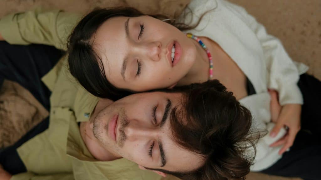 An overhead close-up of a young couple lying on sand with their eyes closed, the woman resting her head on the man's chest.