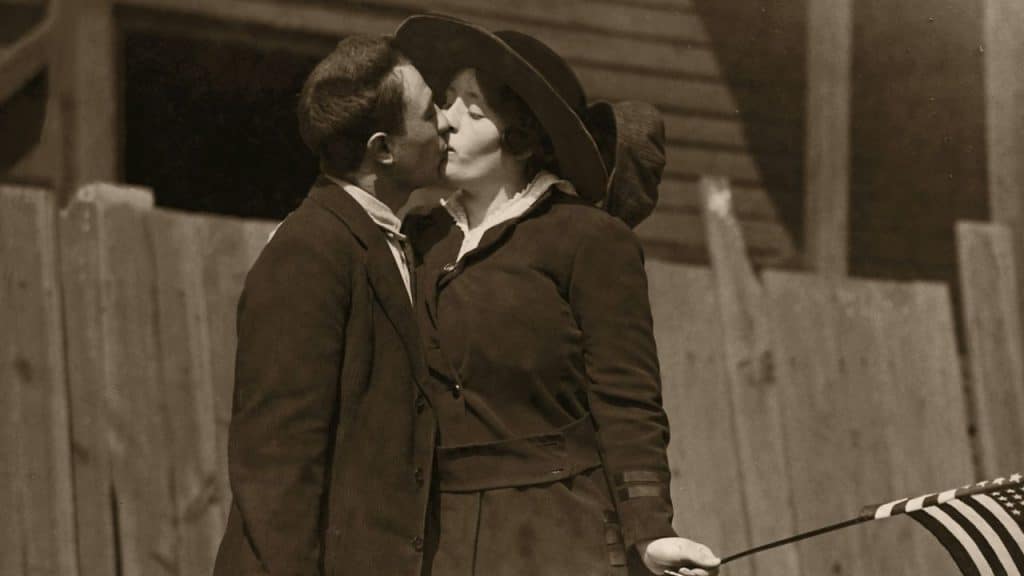A sepia-toned photo of a man and woman kissing outdoors, with the woman holding a small American flag and wearing a wide-brimmed hat.