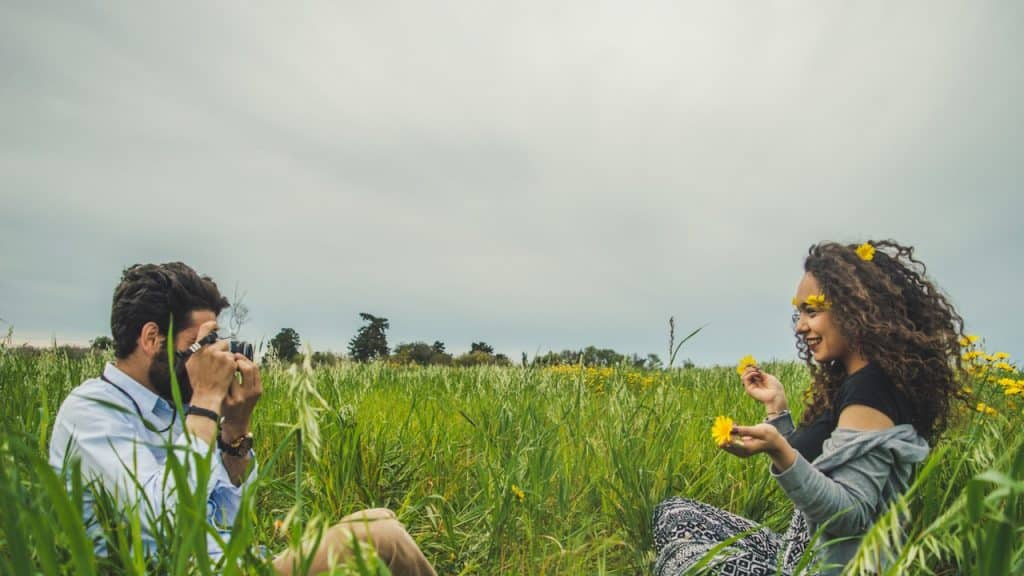 A man taking a photo of a woman sitting in a green field.
