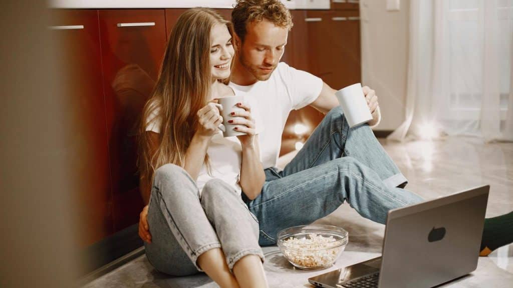 A couple sitting on the floor watching something on a laptop with popcorn and mugs.