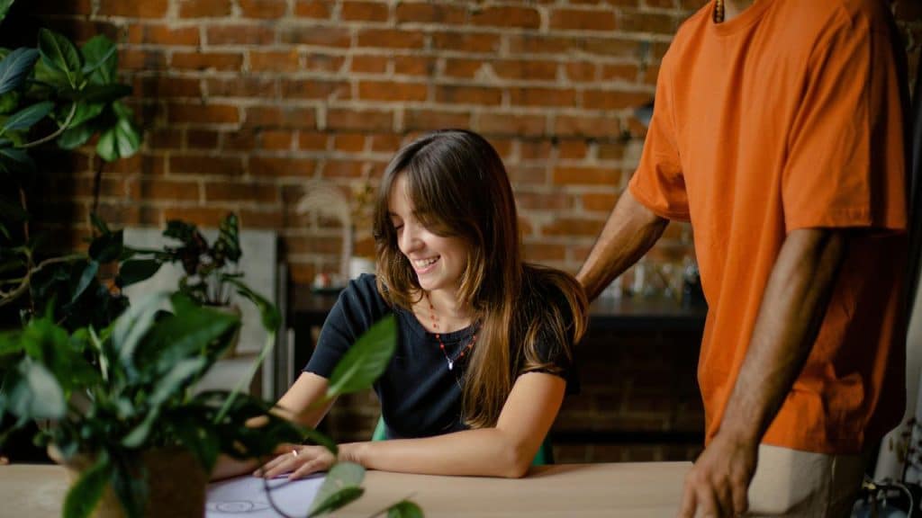 A woman smiling while writing at a table with someone standing beside her.
