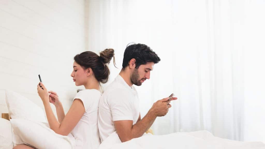 A couple sitting on a bed back-to-back and using cellphones.