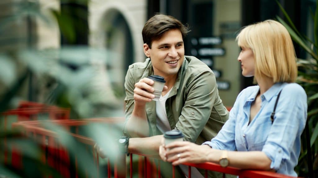 A smiling man and woman stand talking over a red railing, holding coffee cups.