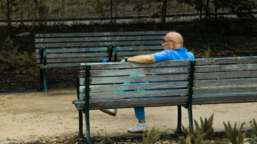 An elderly, bald man in a blue shirt sits alone on a wooden park bench.